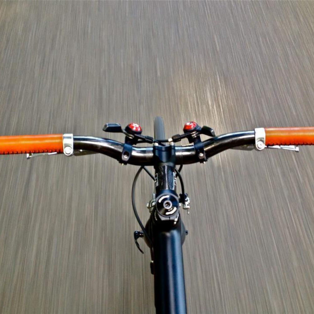 Customer photo looking down on handlebars while riding a bicycle. The road is blurry from motion, the bicycle is dark blue and the leather city handlebar grips are honey color with white thread stitching.