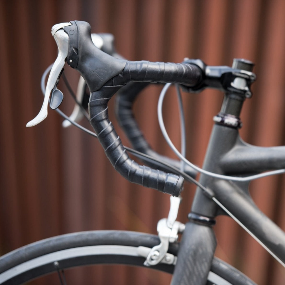 Profile view of the front half of a black bicycle showing drop bars in the center wrapped in coiled black leather bar tape against a rusty corrugated background.