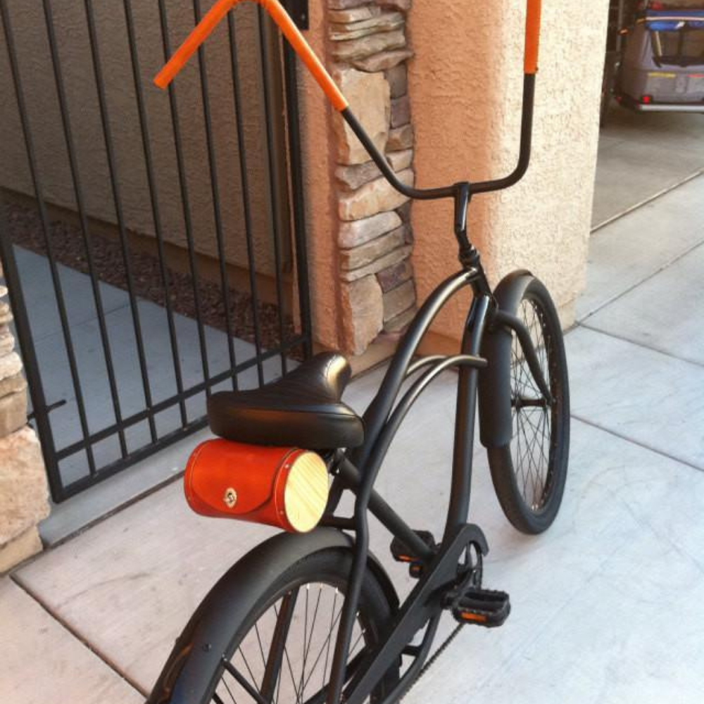 Customer photo of an all-black low rider bicycle with tall monkey handlebars. The handlebars are wrapped in honey sew-on leather bar wraps and there is also a Walnut honey seat barrel bag on the saddle. 