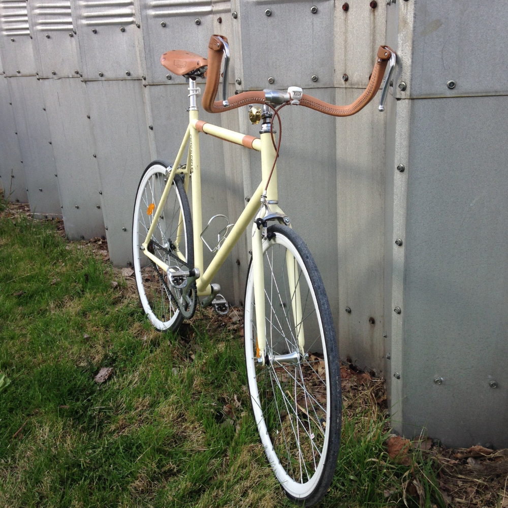 Customer photo of a light yellow bicycle with a natural Brooks saddle and matching Natural sew-on leather bar wraps, which can be ordered custom. The bicycle is against an industrial steel backdrop on a grass patch.