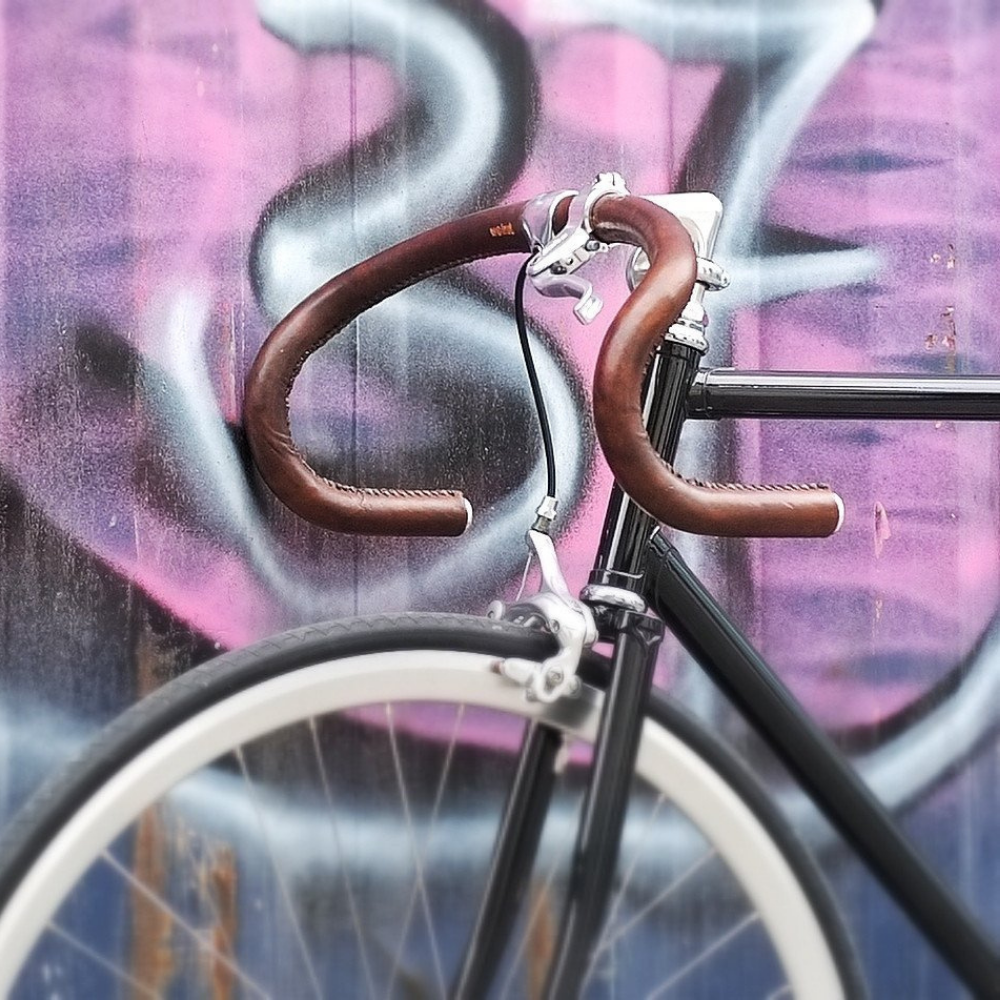 Angled view of bicycle handlebars with dark brown sew-on leather bar wraps. The bicycle is black and up against a purple graffiti wall as a backdrop.