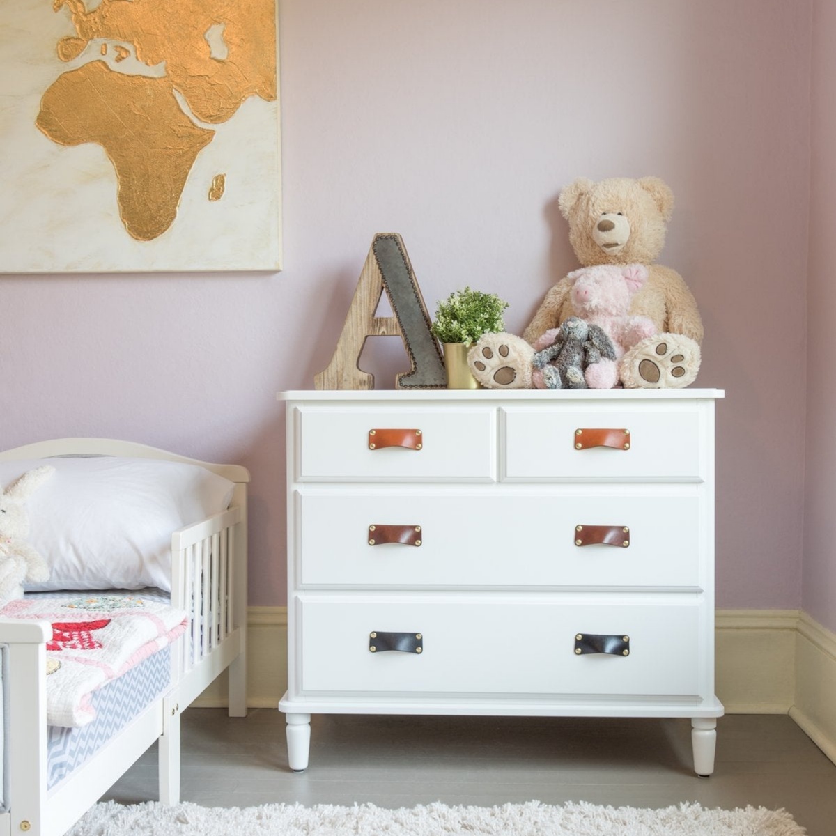 Children&#39;s nursery with natural leather bin pulls on clean white furniture. A large teddy bear sits on top of the chest of drawers and the walls are painted a pastel purple with gold accents. 