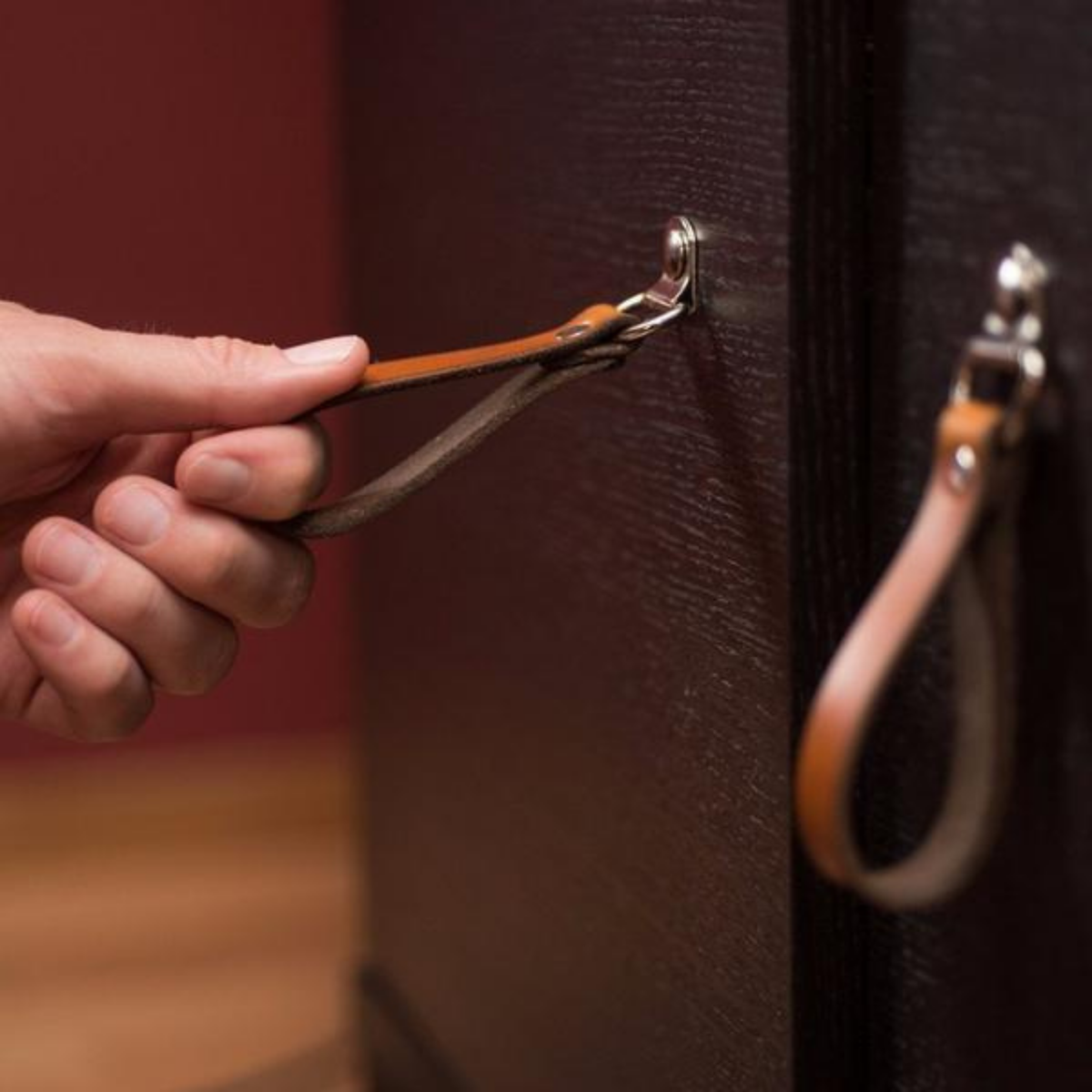 Man&#39;s hand reaching for a cognac colored leather hinged handle on a dark brown cabinet door. 