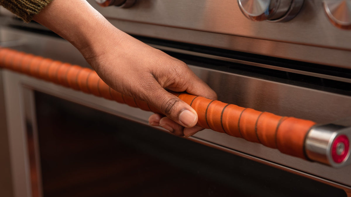 Close-up of a leather-wrapped oven handle with a hand reaching down to open the door. 
