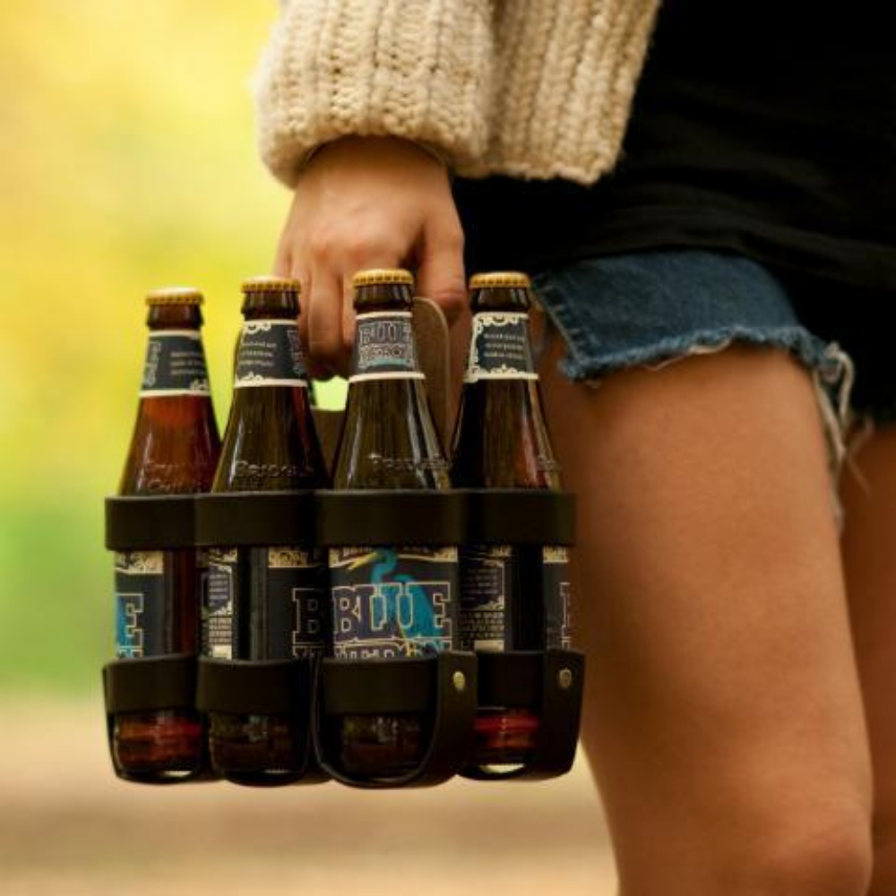 A woman holding a 6-pack of beer in a handsome leather and wood reusable 6-pack carrier