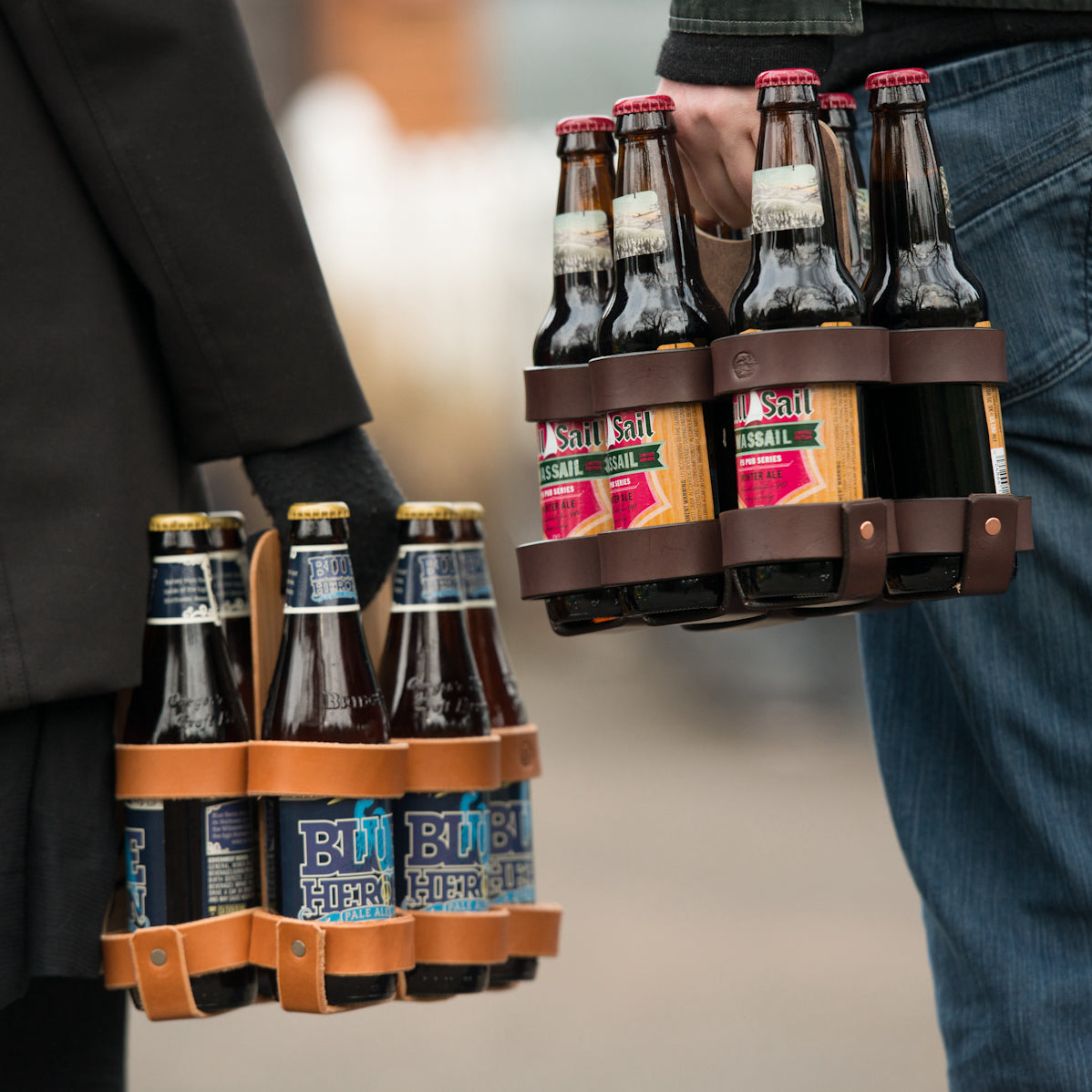 A man and a woman each holding a 6-pack of beer in a handsome leather and wood reusable 6-pack carrier, with different beers and different leather colors. 