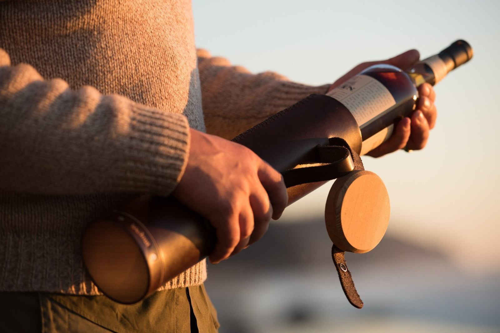A man in a tweed sweater is pulling a bottle of Oban scotch whisky out of a handcrafted leather tube with an attached wood lid at sunset on the beach