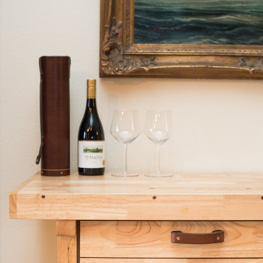 A leather bottle case next to a bottle of wine that fits inside with two wine glasses on a sideboard / buffet table that also has Tilikum leather drawer handles on it. 