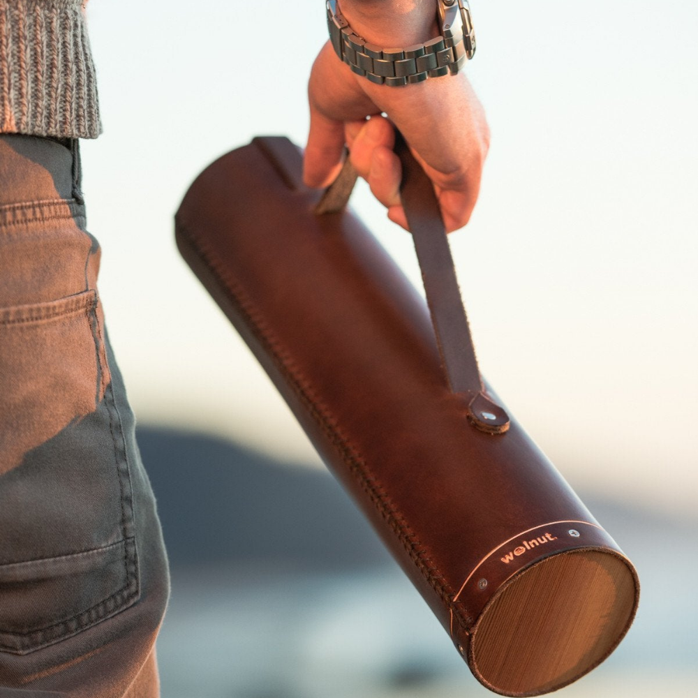 Man wearing a woolen sweater and a chunky watch holding a closed dark brown leather whiskey case by the handle strap. The bottom of the case is cedar wood and he is walking on the Oregon Coast beach.