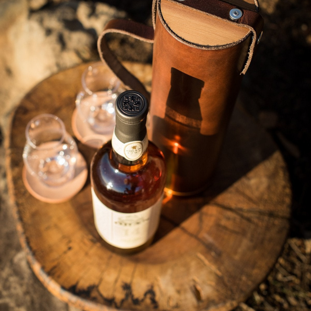 Top-down photo of a still life scene, showing a bottle of Scotch whisky, two Glencairn glasses on leather coasters, and a dark brown leather whiskey bottle case on a natural wood cutting board at the beach. 