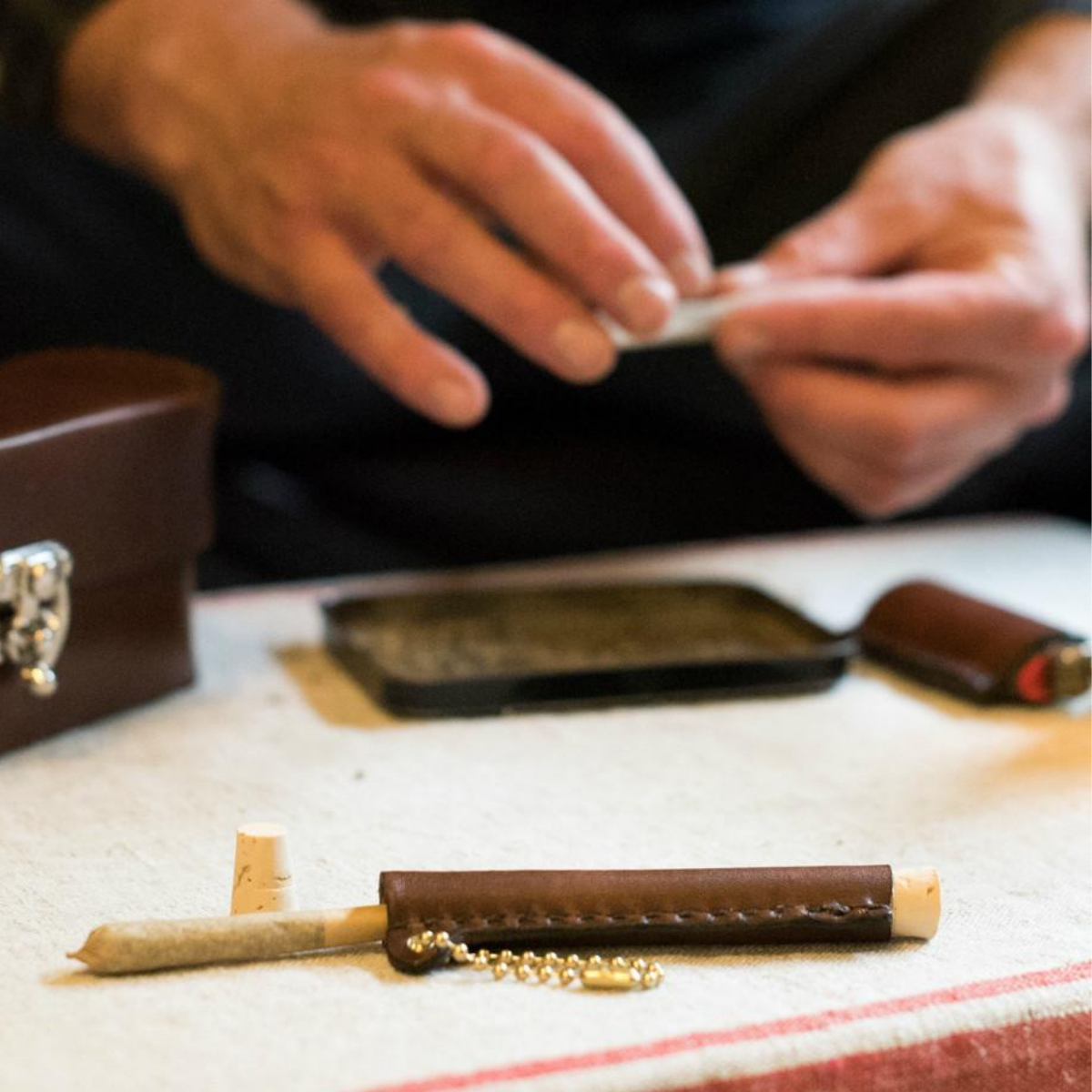 Man rolling a cigarette next to a leather &quot;Classy Stash&quot; box and leather lighter holder
