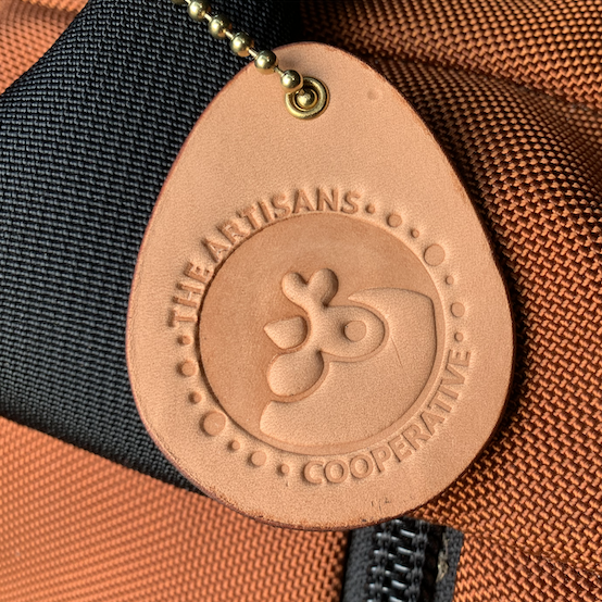 Close-up of the egg-shaped leather fundraiser keychain for Artisans Cooperative attached to a orange luggage bag with brass ball chain as a luggage identifier tag