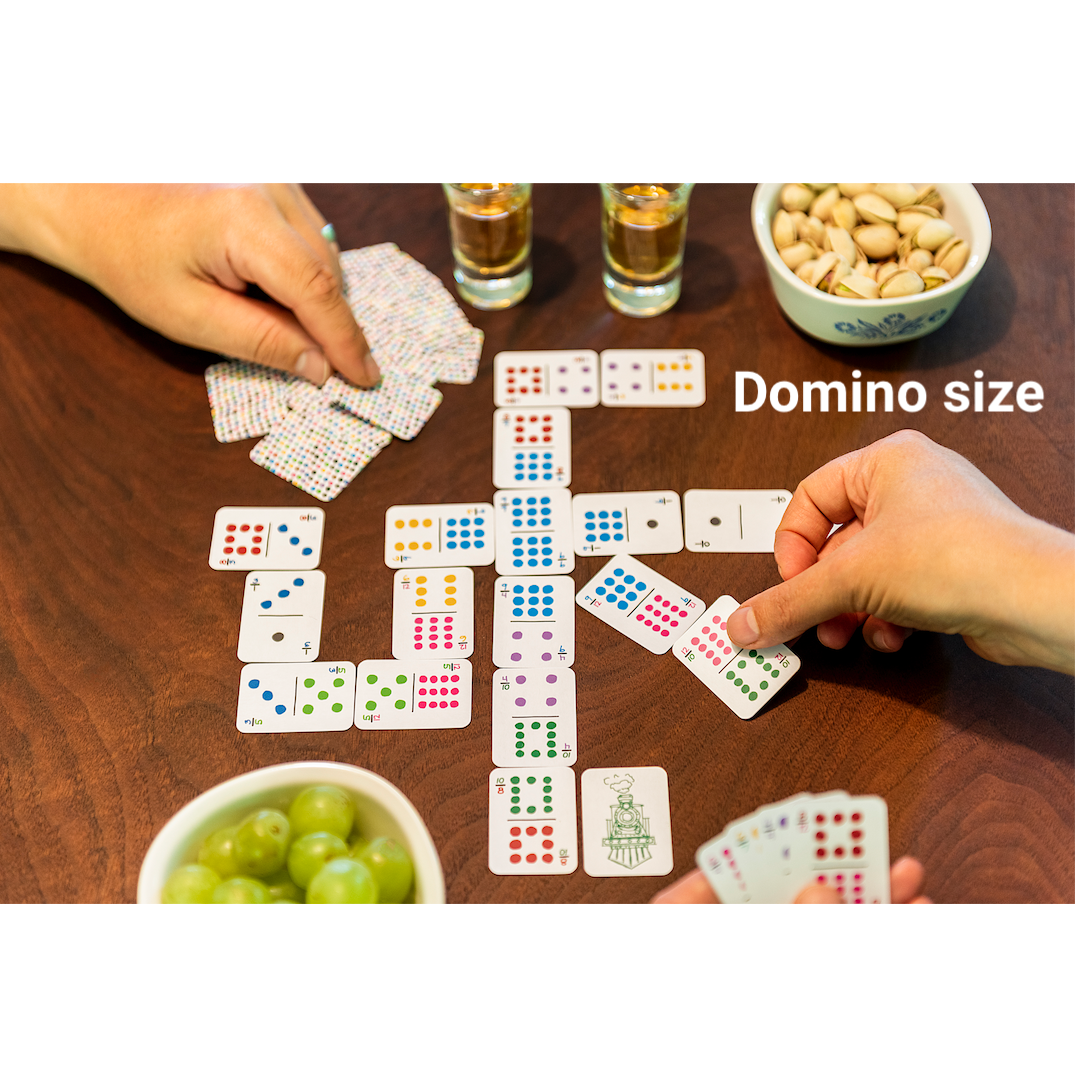 Photo of domino-sized mini playing cards (one quarter the size of standard playing cards) on a table with props for scale: two hands playing, a bowl of grapes and pistachios, a shot of whiskey, and the words &quot;Domino size&quot; on top of the photo