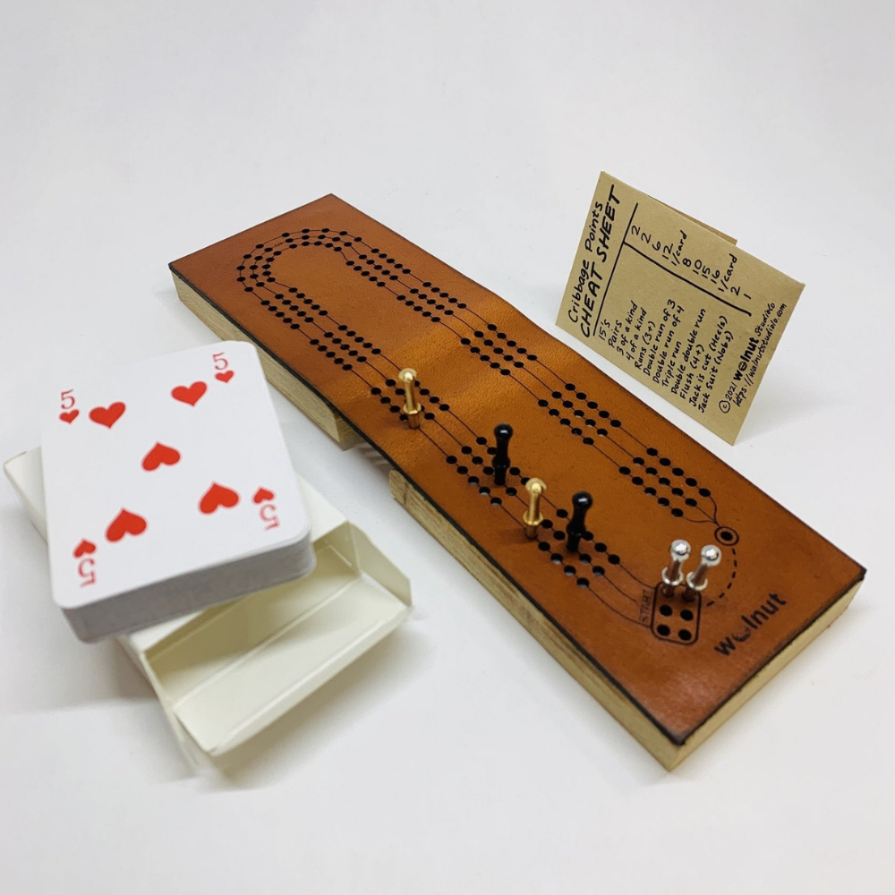 White background photo showing the Mini Deluxe leather folding travel cribbage board with all its included parts: a half-sized deck of playing cards, a coin envelope for storing pegs, and three sets of cast metal pegs. 