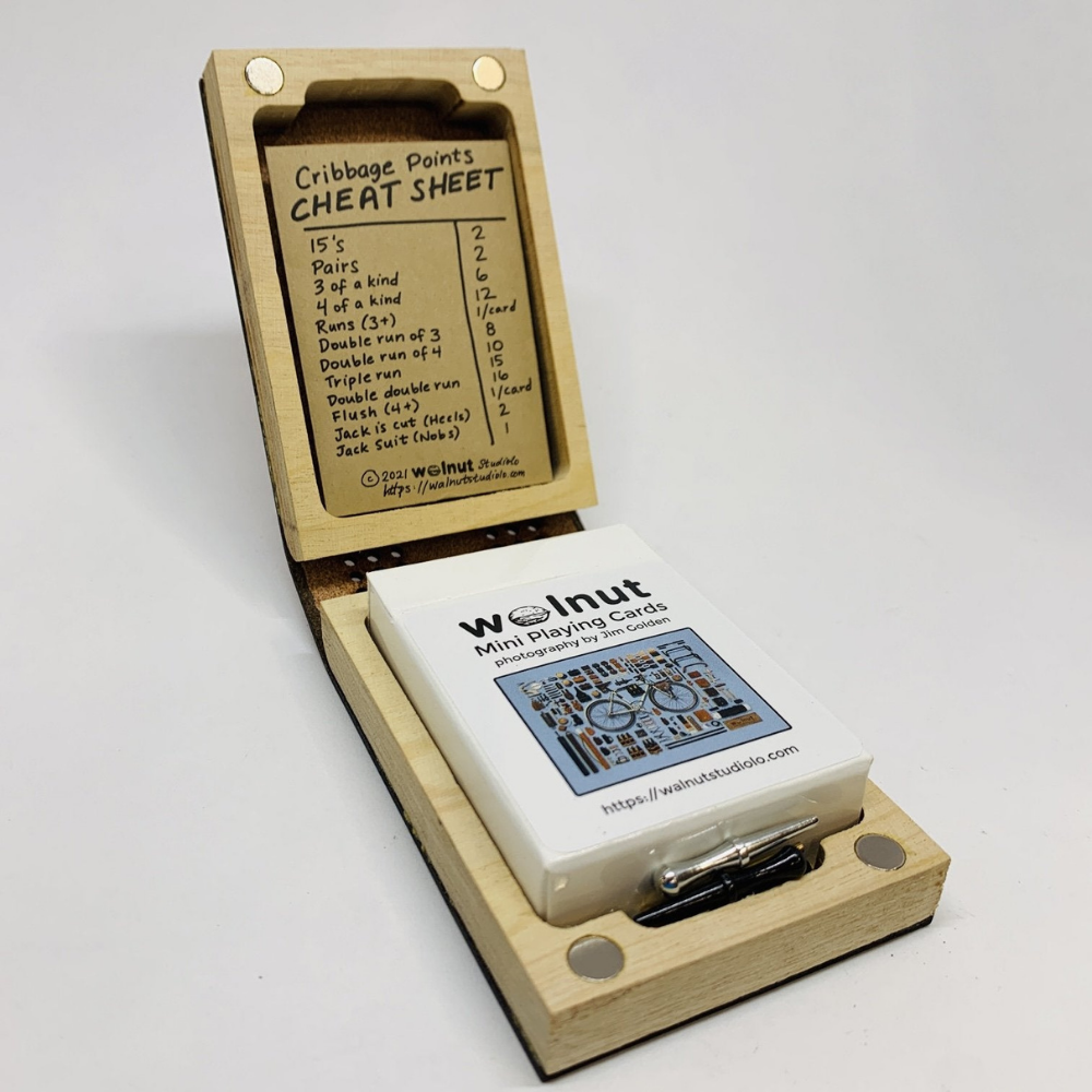 Unfolded leather mini deluxe travel cribbage board, showing wood perimeter and interior playing card storage for a half-sized deck of cards. Also shows a small coin envelope that holds three sets of cast metal pegs in silver, black, and brass finish.