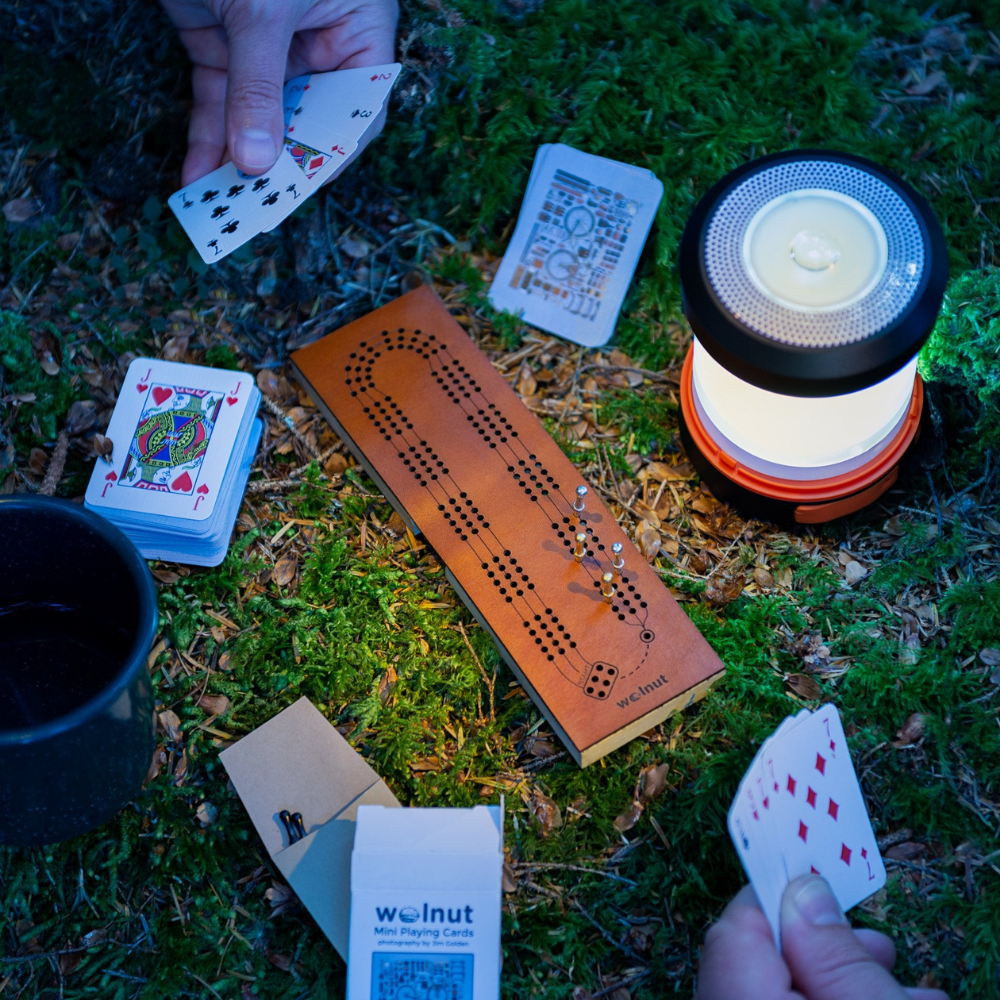 Small folding leather cribbage board being played at night while camping with an LED lantern and half-size mini playing cards for backpacking. 