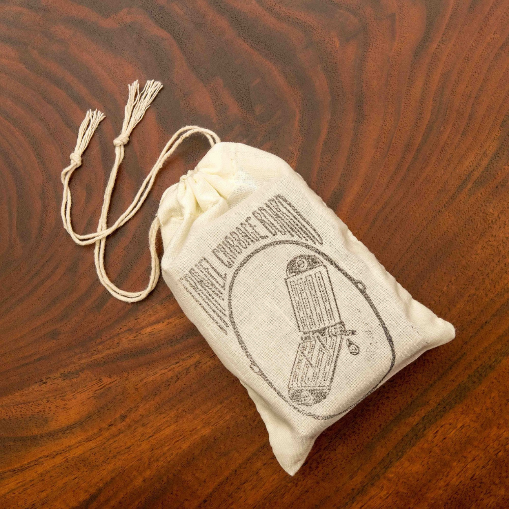 Gift Set Muslin Carrying Pouch on walnut table. The muslin pouch has a hand-drawn illustration of the Travel Cribbage Board with the name of it stamped on the surface. 