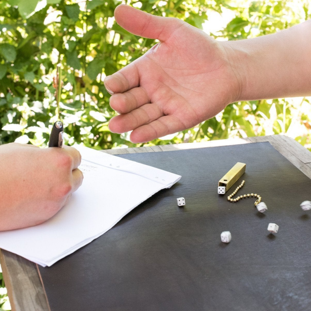 Two people playing dice on a summery day with green bushes in the background. One person is rolling six small travel dice and the other person is keeping score with a pen and paper. The brass storage tube is on the table.